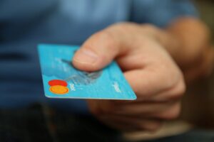 A person in a blue shirt offers a Samsung credit card with focus on the card, which features a silver hologram and a chip. The background is blurred.