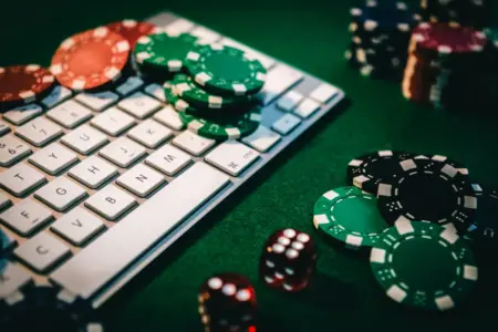 A close-up view of a white keyboard with casino chips and dice scattered around it on a green surface, suggesting online gambling enabled by technological advances.