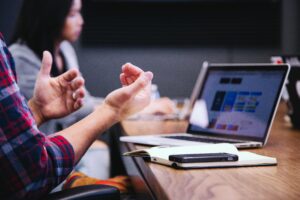 A close-up of a person's hands gesturing during a business meeting with a laptop displaying marketing strategies and a colleague in the background.