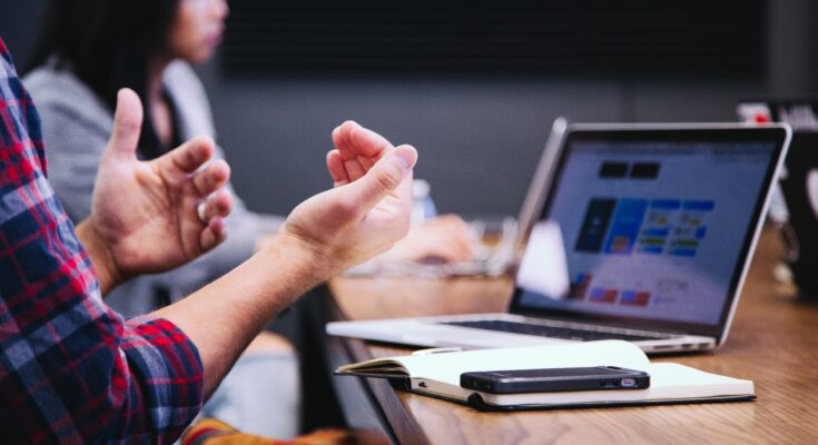 A close-up of a person's hands gesturing during a business meeting with a laptop displaying marketing strategies and a colleague in the background.