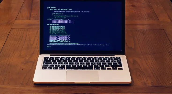 A laptop open on a wooden table displaying lines of secured code on its screen, illuminated in a room with soft lighting.