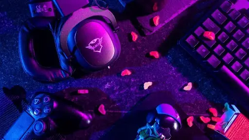 A gaming setup featuring an esports keyboard, headphones, a controller, and a mouse, all illuminated in blue and purple lights. Scattered around are small red heart-shaped candies.