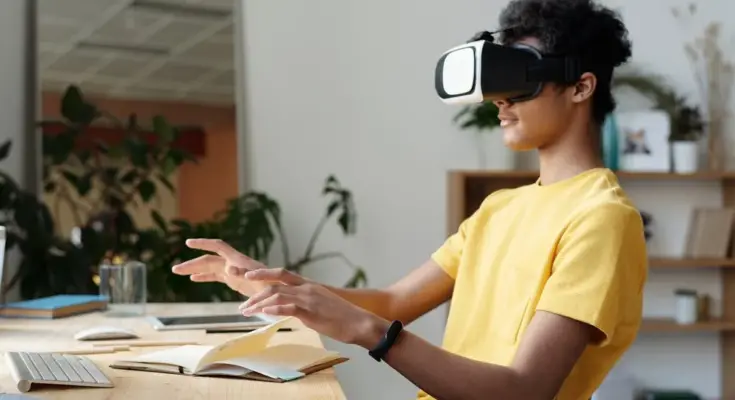 A person wearing a VR headset sits at a desk, interacting with an unseen virtual environment, surrounded by a laptop, books, and a technology trends office setting.