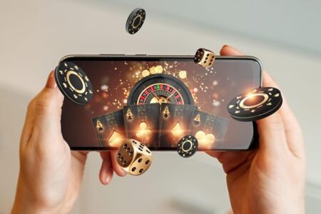 A person holds a smartphone displaying 3D graphics of a roulette wheel, playing cards, casino chips, and dice floating out of the screen, symbolizing online gaming.
