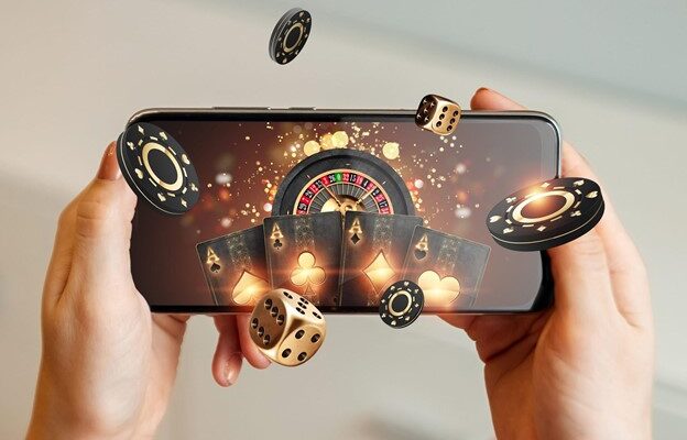 A person holds a smartphone displaying 3D graphics of a roulette wheel, playing cards, casino chips, and dice floating out of the screen, symbolizing online gaming.