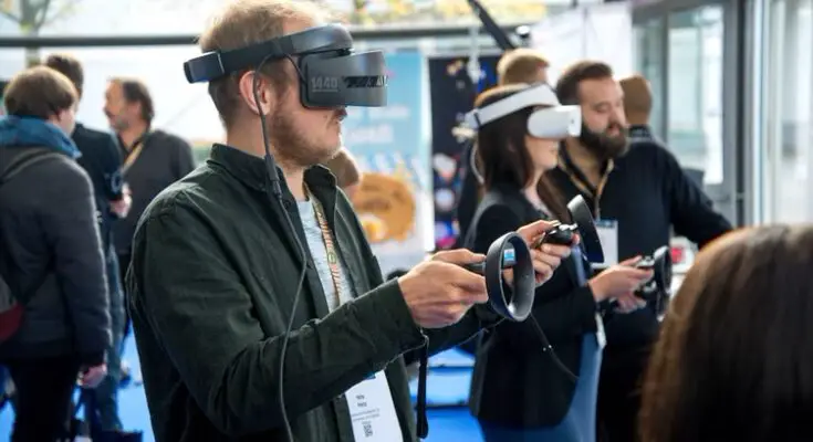 A man wearing a virtual reality headset and holding controllers stands at a tech expo, showcasing the growing industries of VR technology, with other attendees in VR gear around him.