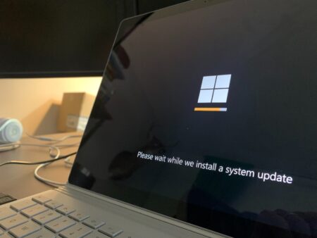 A laptop screen displaying a windows system patch progress with the text "please wait while we install a system update" on a dark background.