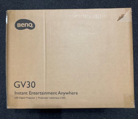 A BenQ GV30 cardboard box on a floor, labeled with "instant entertainment anywhere, portable projector." The box is closed and appears new.