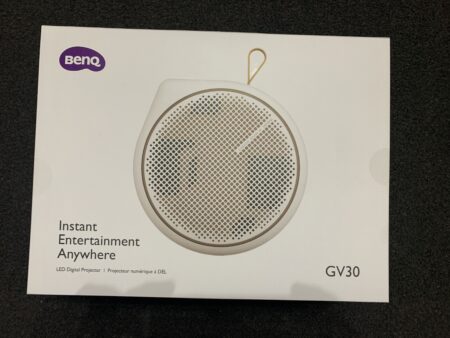 A BenQ GV30 portable projector packaging box, featuring an image of the round, cream-colored projector with a grid speaker design on top. The box highlights the product's feature of offering "instant entertainment