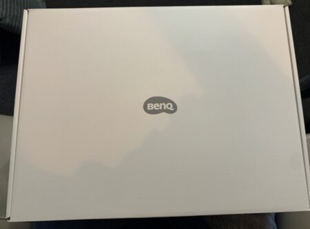 A closed, plain white box with the "benq" logo at the center, placed on a dark fabric surface, containing a 2.1 channel Bluetooth speaker, with light glare visible on the