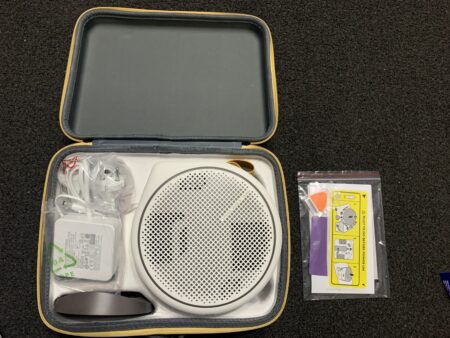 Opened travel case on the floor displaying a 2.1 channel Bluetooth speaker, a charger, and a pack of batteries. The case is neatly organized, with the items arranged on a black background.