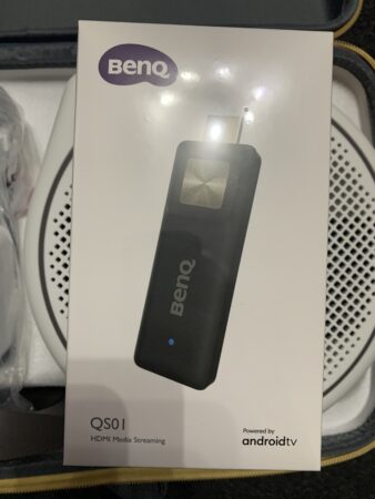 A benq q501 hdmi media streaming device powered by android TV, packaged in a plastic cover, displayed on a patterned background.