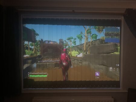 A video game screen showing a character in a pink outfit viewed from the back, displayed on a ceiling projection seen through a window blind in a dimly lit room.