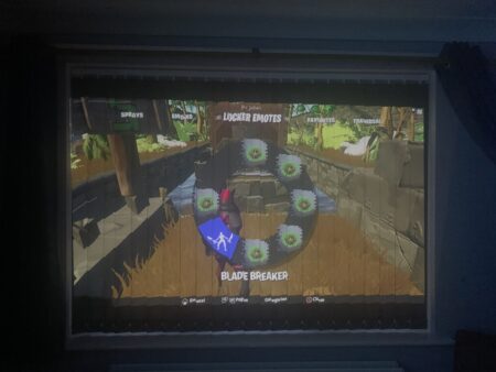 A BenQ GV30 portable projector displaying a video game interface on a ceiling, viewed through a window frame in a dark room. The game shows a character in a virtual environment with a menu overlay for
