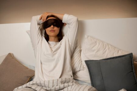 A woman sits in bed with a sleep mask pushed up onto her forehead, surrounded by pillows, wearing a white shirt, holding an iOS device, appearing relaxed or getting ready to sleep.