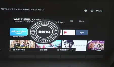 A computer monitor displaying a selection screen with various streaming service apps, including Prime Video and others in Japanese, on a graphic interface resembling a wheel. Time display reads 9:03. The setup includes