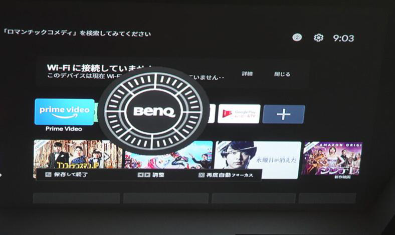 A computer monitor displaying a selection screen with various streaming service apps, including Prime Video and others in Japanese, on a graphic interface resembling a wheel. Time display reads 9:03. The setup includes