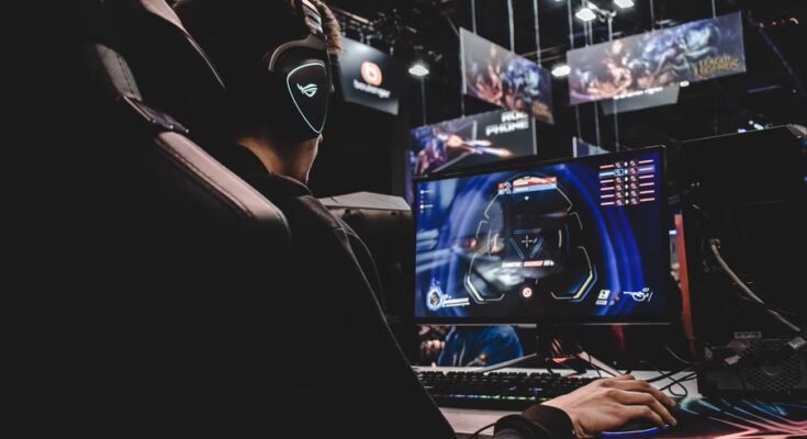 A person with headphones is intensely focused on a gaming session on a computer, surrounded by the vibrant lights and screens of a gaming event.