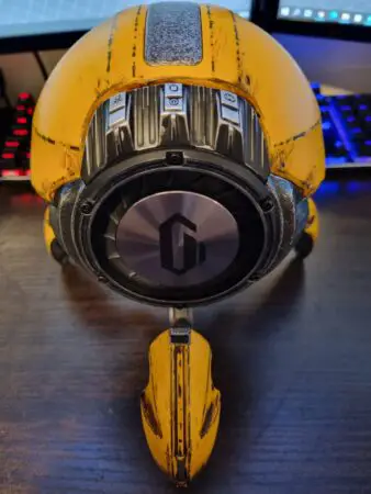 A close-up image of a weathered yellow motorcycle helmet with black accents, positioned on a desk. The helmet features a reflective, dark visor, Bluetooth speaker integration, and a distinctive, streamlined design