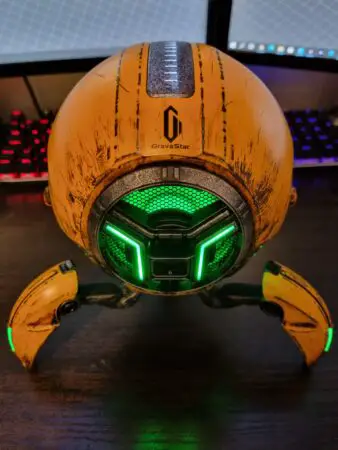 A futuristic orange and black GravaStar robot-shaped Bluetooth speaker with glowing green lights, set against a computer monitor background.