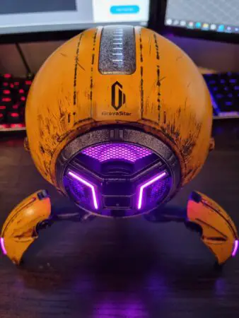 A futuristic, orange Bluetooth speaker with purple lighting, branded "GravaStar," set against a backdrop of a computer keyboard with rainbow-colored backlighting.