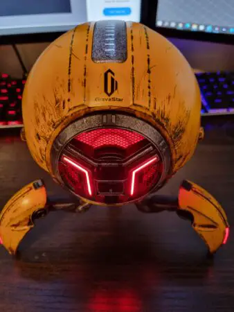 A GravaStar futuristic-looking yellow and black Bluetooth robot-style speaker with red illuminated elements, positioned on a desk with a computer in the background.