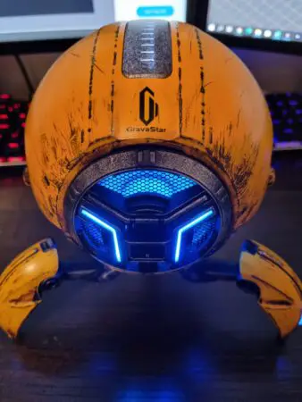A futuristic orange Bluetooth speaker branded "gravastar" with spider-like legs, featuring bright blue LED lights on the face, set against a backdrop of a computer keyboard.