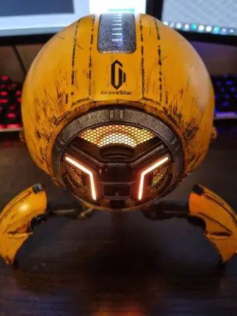 A close-up photo of a GravaStar robotic-themed Bluetooth speaker with a distinctive yellow and black design, featuring illuminated orange eyes and a mesh grille.