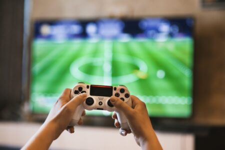 Person playing a gaming soccer video game, holding a white gaming controller with screen showing a soccer match in the background.