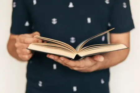A gamer in a dark shirt adorned with small anchor prints holds an open book with pages slightly flipping, against a neutral background.