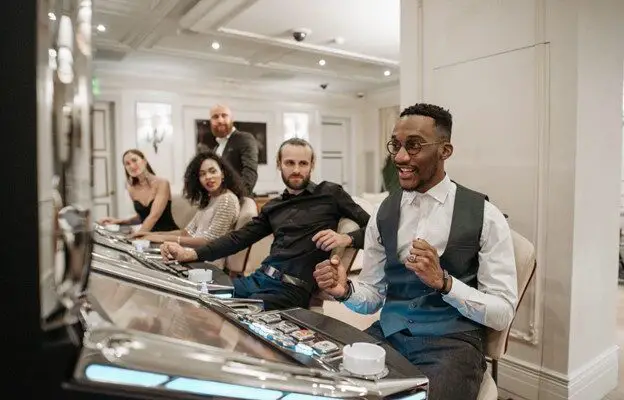 Group of five people in formal attire sitting at a bar, with one man in the foreground smiling and gesturing excitedly about casino tips.