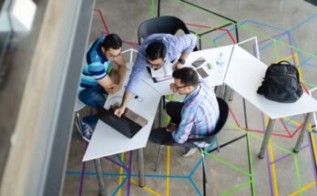 Three people around a geometric table in a modern workspace, engaged in a discussion about DDI with laptops and notebooks, viewed from above.