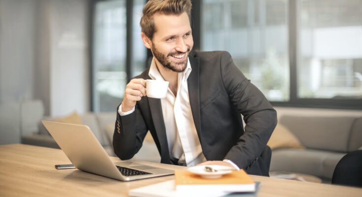 man holding white teacup in front of gray laptop