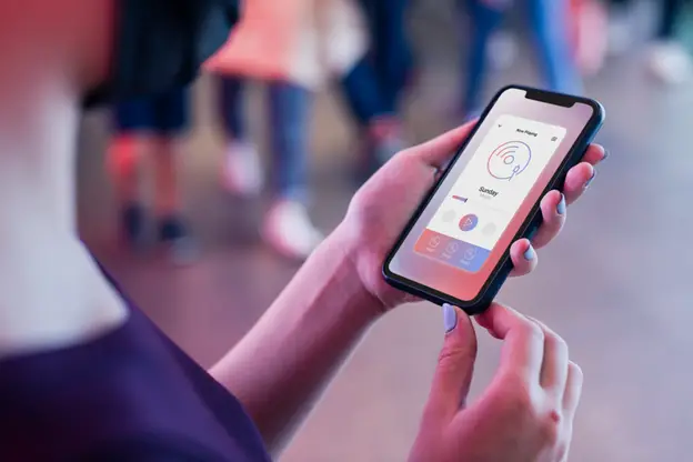 A person holds a smartphone displaying a music app development interface, with focus on the screen and blurred people moving in the background.
