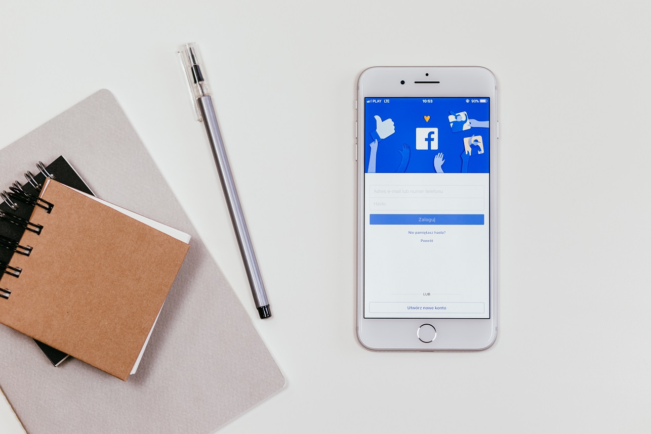 An iPhone displaying the Facebook Ads login page, resting on a white surface next to a closed notebook and a pen. The scene suggests a modern, connected lifestyle or a moment of digital interaction.