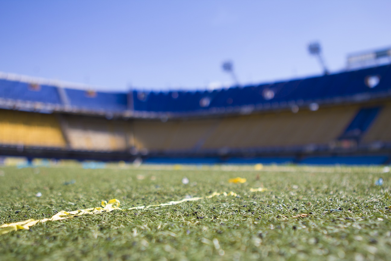 Close-up view of a soccer field's grass with blurred stadium seats in the background, under a clear blue sky, ideal for making sports bets.