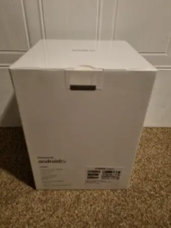 A white XGIMI Halo+ projector box packaging standing upright on a carpeted floor against a light-colored door, showing the front side with product logos and information.