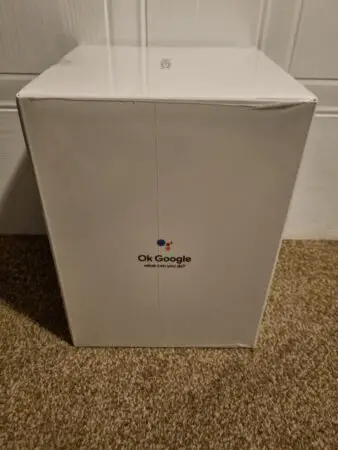 A large box stands upright against a wall, labeled with the "ok google" logo and the phrase "what can you ask?" on its side. The box appears to be a product packaging for the