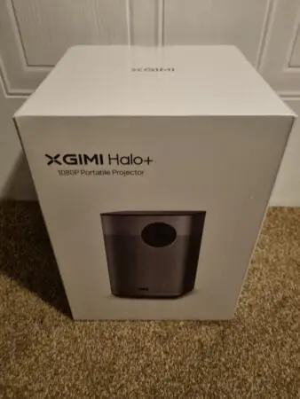 A sealed box of a XGIMI Halo+ 1080p portable projector standing upright against a door, prominently displaying the product image and XGIMI logo on the front.