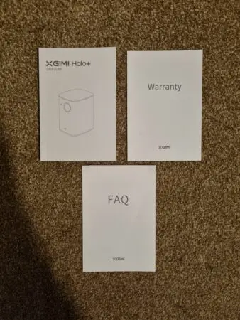 Three documents on a carpet: top left is an XGIMI Halo+ user guide, top right is a warranty paper, and bottom is an FAQ sheet, all branded with XGIMI