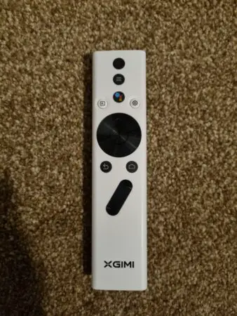 A white XGIMI Halo+ brand remote control is lying on a textured tan carpet. The remote features a black navigation pad, multiple control buttons in various colors, and the logo near the bottom