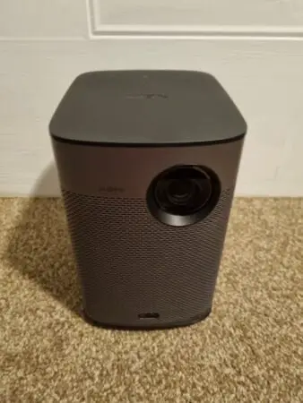 A black XGIMI Halo+ portable projector on a carpeted floor against a white wall background. The projector has a prominent lens on the upper front and mesh texture around its body.