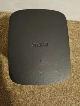 A XGIMI Halo+ projector displayed from a top-down view, showing a dark gray matte surface with the brand logo and control symbols (power, volume up, and volume down).