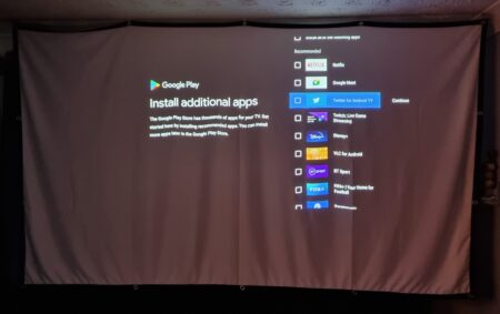 A projected image, displayed using a portable XGIMI projector, shows the Google Play homepage on a screen, featuring a list of recommended apps on the right side.