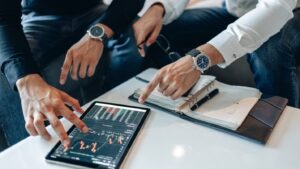 Two individuals analyzing financial data on a tablet, with documents and a notepad on a white table. Both are wearing wristwatches and stylish clothing, focusing intently on the screen as part of their