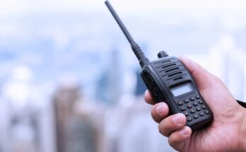 A person's hand holding a two-way radio against a blurred city skyline in the background.