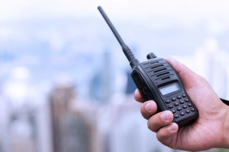 A person's hand holding a two-way radio against a blurred city skyline in the background.