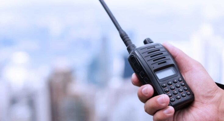 A person's hand holding a two-way radio against a blurred city skyline in the background.