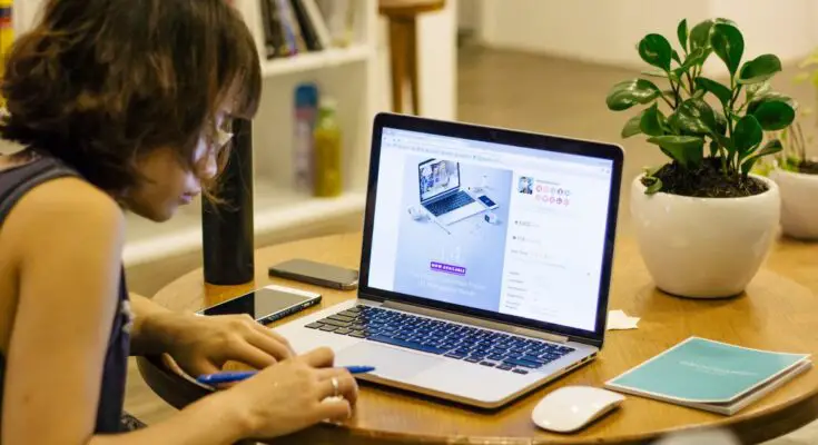 A woman sitting at a wooden table working on a laptop with knowledge base software, with a smartphone, notebook, and potted plant beside her.