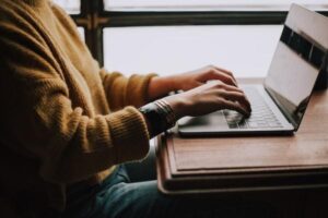 Person in a yellow sweater searching for the best car insurance on a laptop at a wooden table near a window.